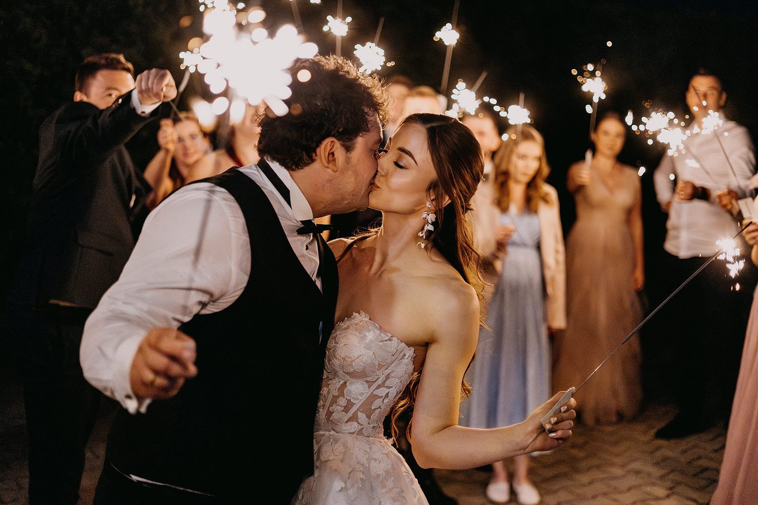 Bride and groom kissing surrounded by wedding guests holding sparklers at a romantic nighttime celebration.