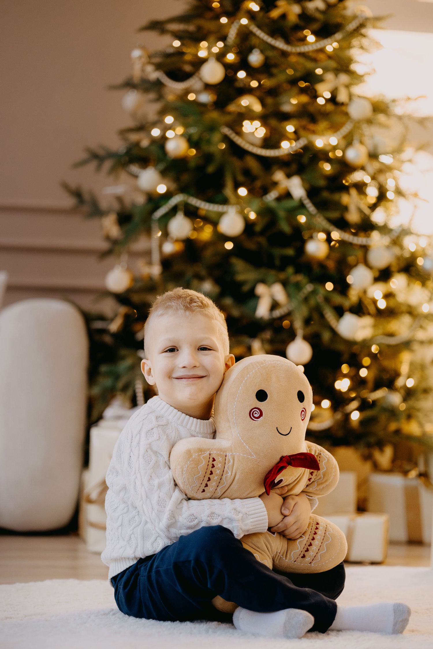 Smiling young boy in a cream knit sweater sits on a white rug hugging a plush gingerbread man in front of a lit Christmas tree.