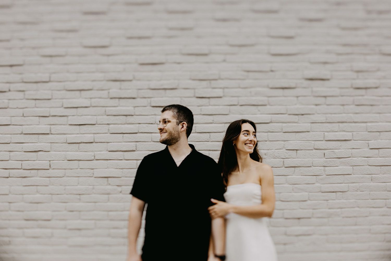 Couple posing in front of a white brick wall, man in black shirt and woman in white dress, smiling outdoors.