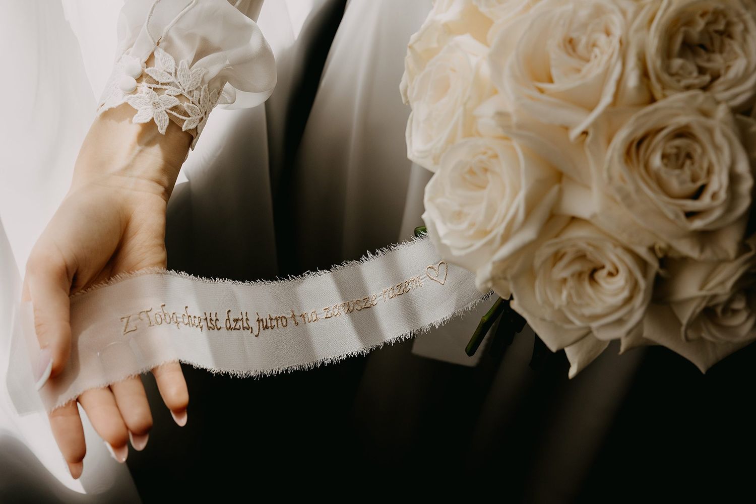 Close-up of bride holding white rose bouquet with ribbon inscription and delicate lace wrist detail.