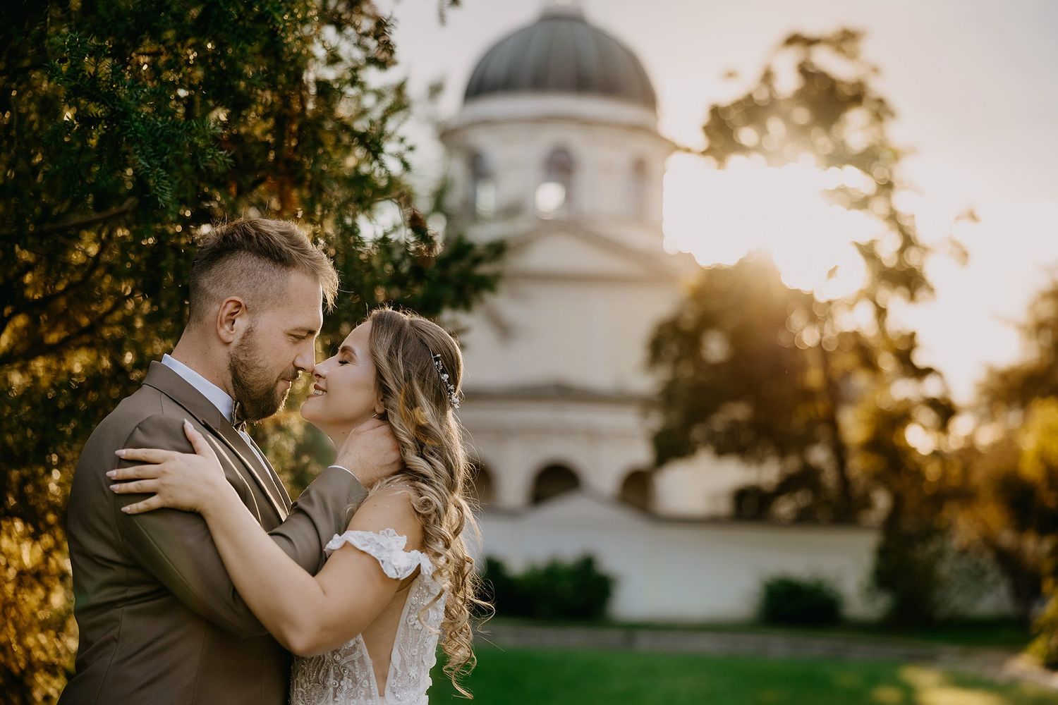 Bride and groom sharing intimate moment near ornate domed building in warm golden sunlight.