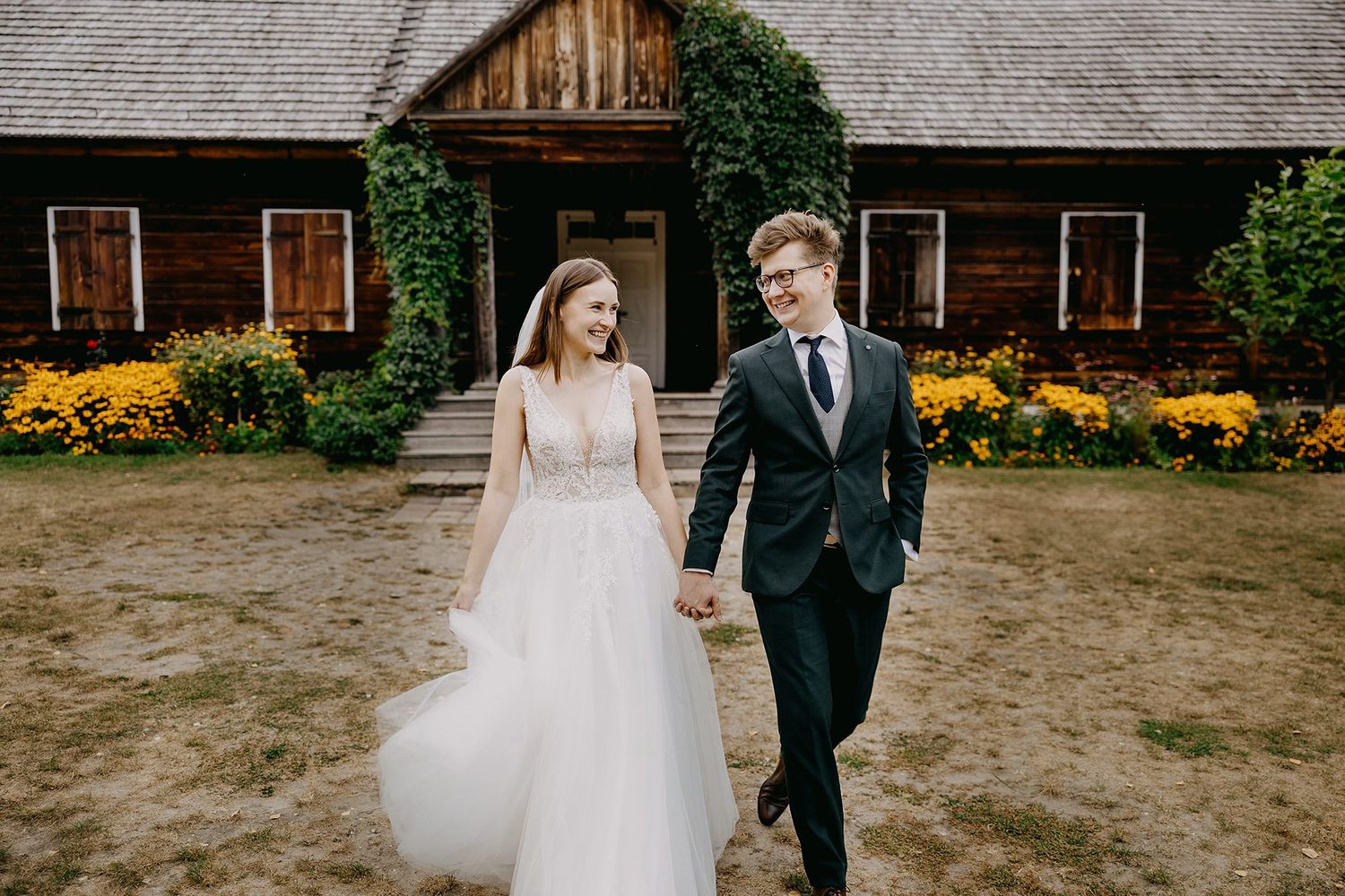 Smiling bride and groom holding hands walking in front of a rustic wooden building with yellow flowers.