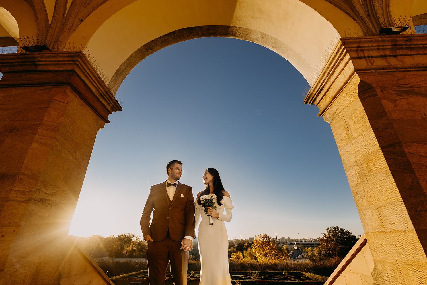 Bride and groom stand beneath stone arch with golden sunlight and blue sky at wedding venue.