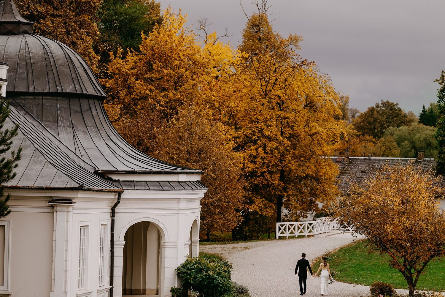 Wedding couple walking along path beside historic white chapel amid autumn golden trees.
