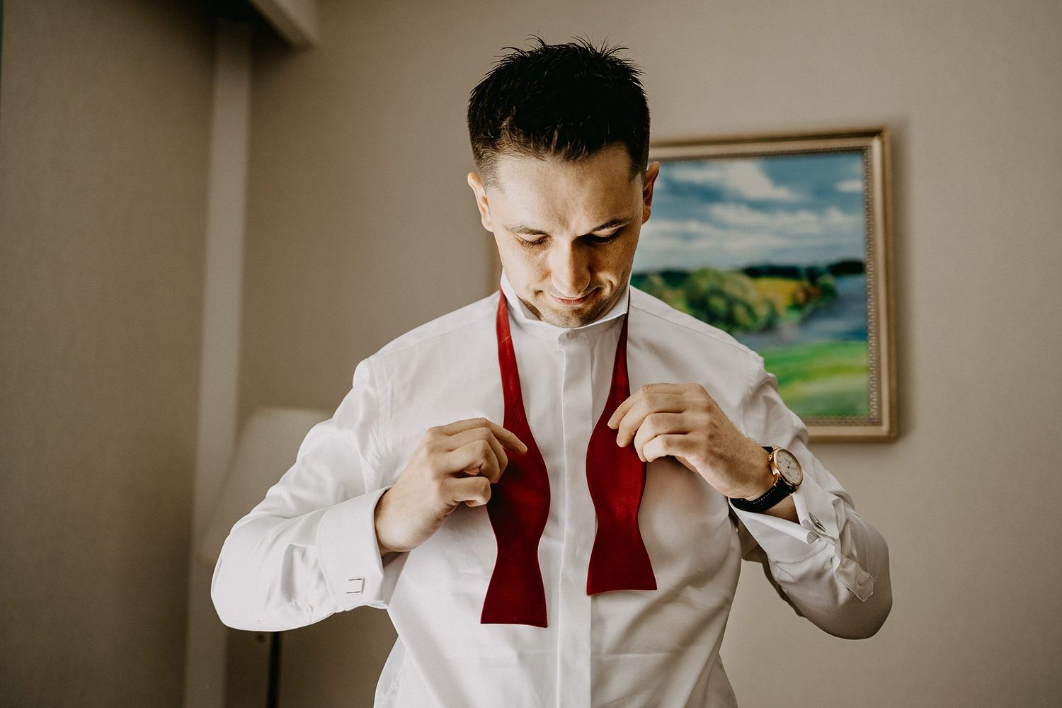 A dapper groom adjusts his red bow tie while getting ready in a well-lit room before his wedding ceremony.