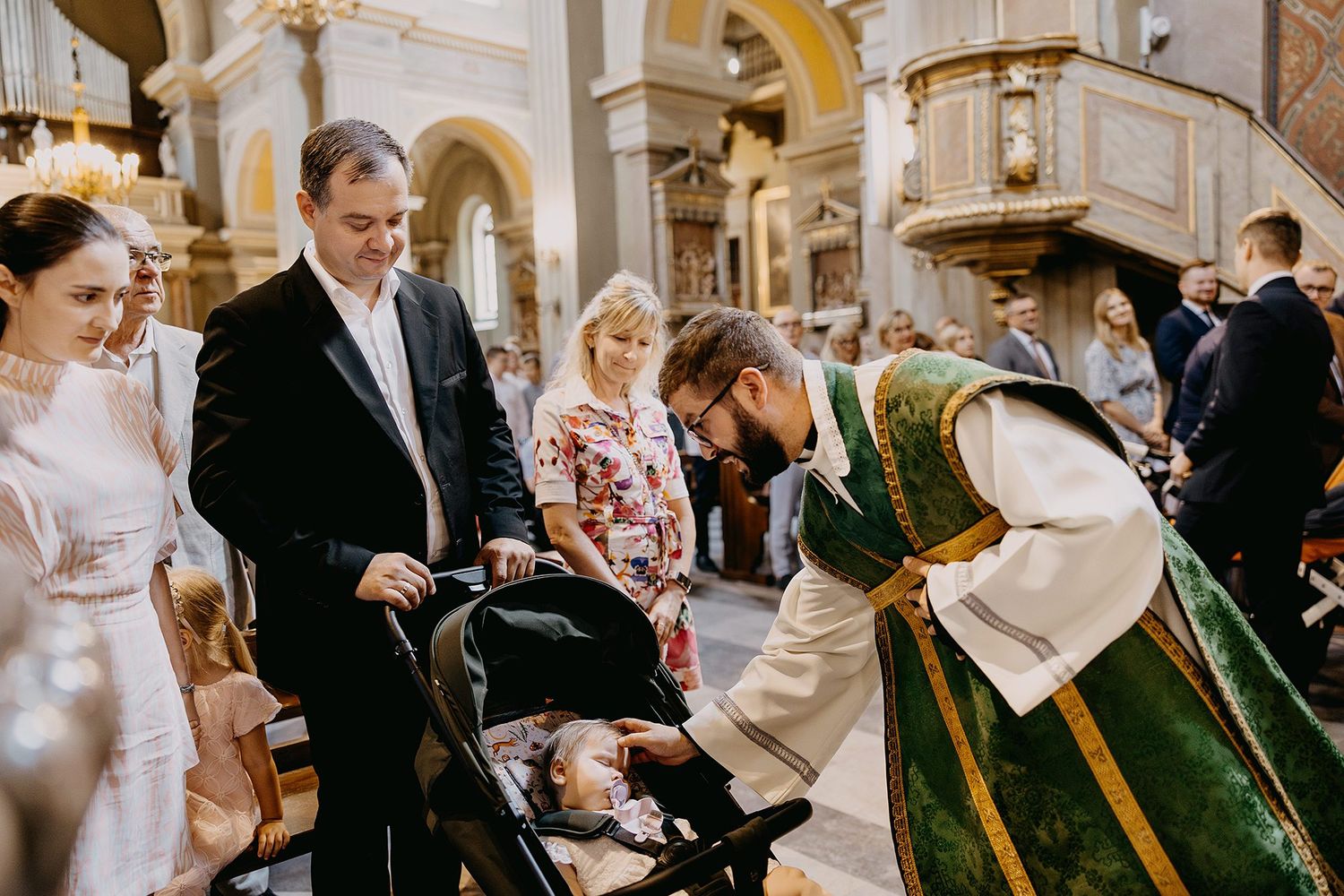 Catholic priest in green vestments bending to bless infant in stroller during church baptism ceremony.