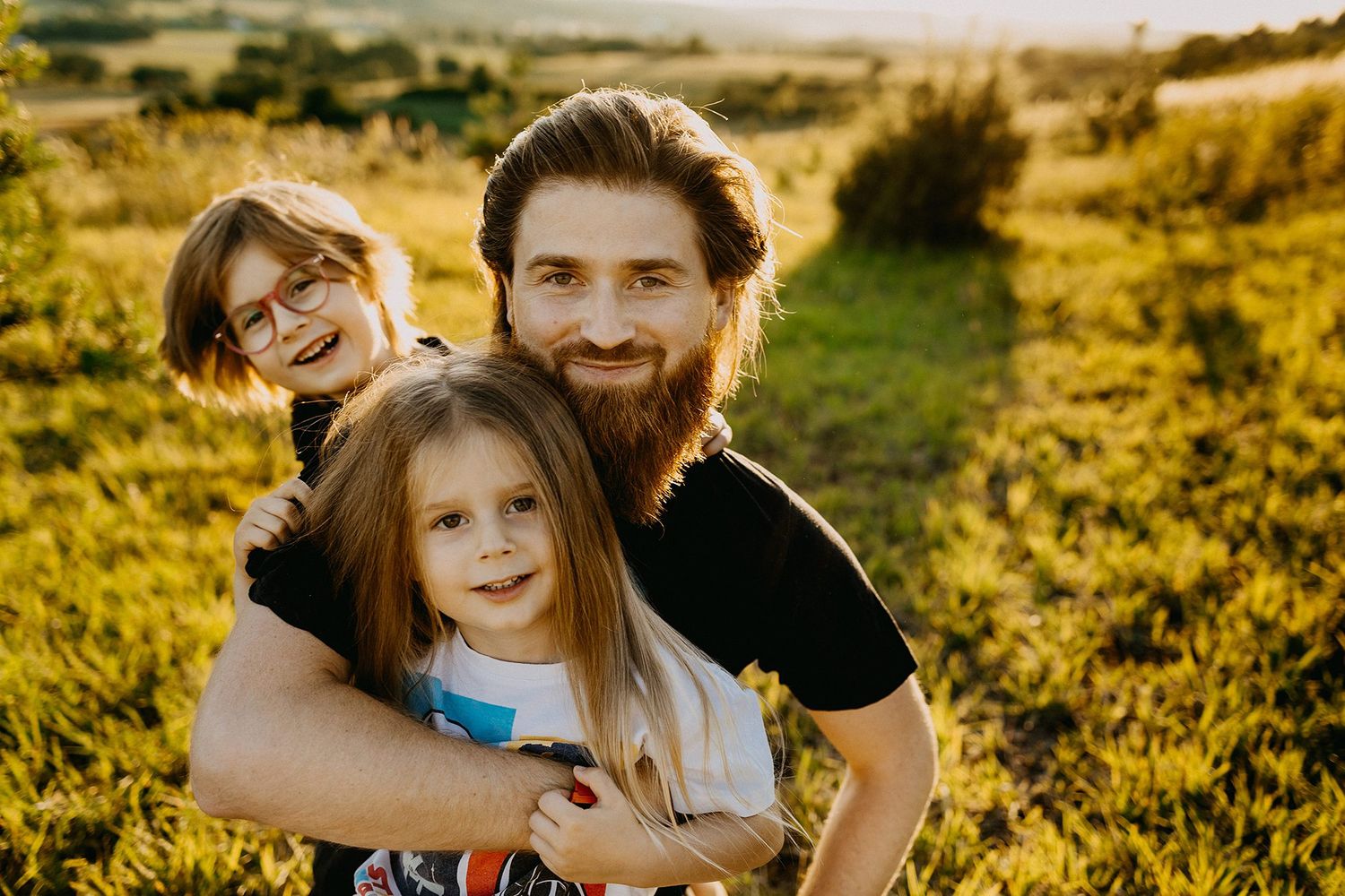 Bearded father and mother with glasses hugging young daughter in a sunny green countryside field.