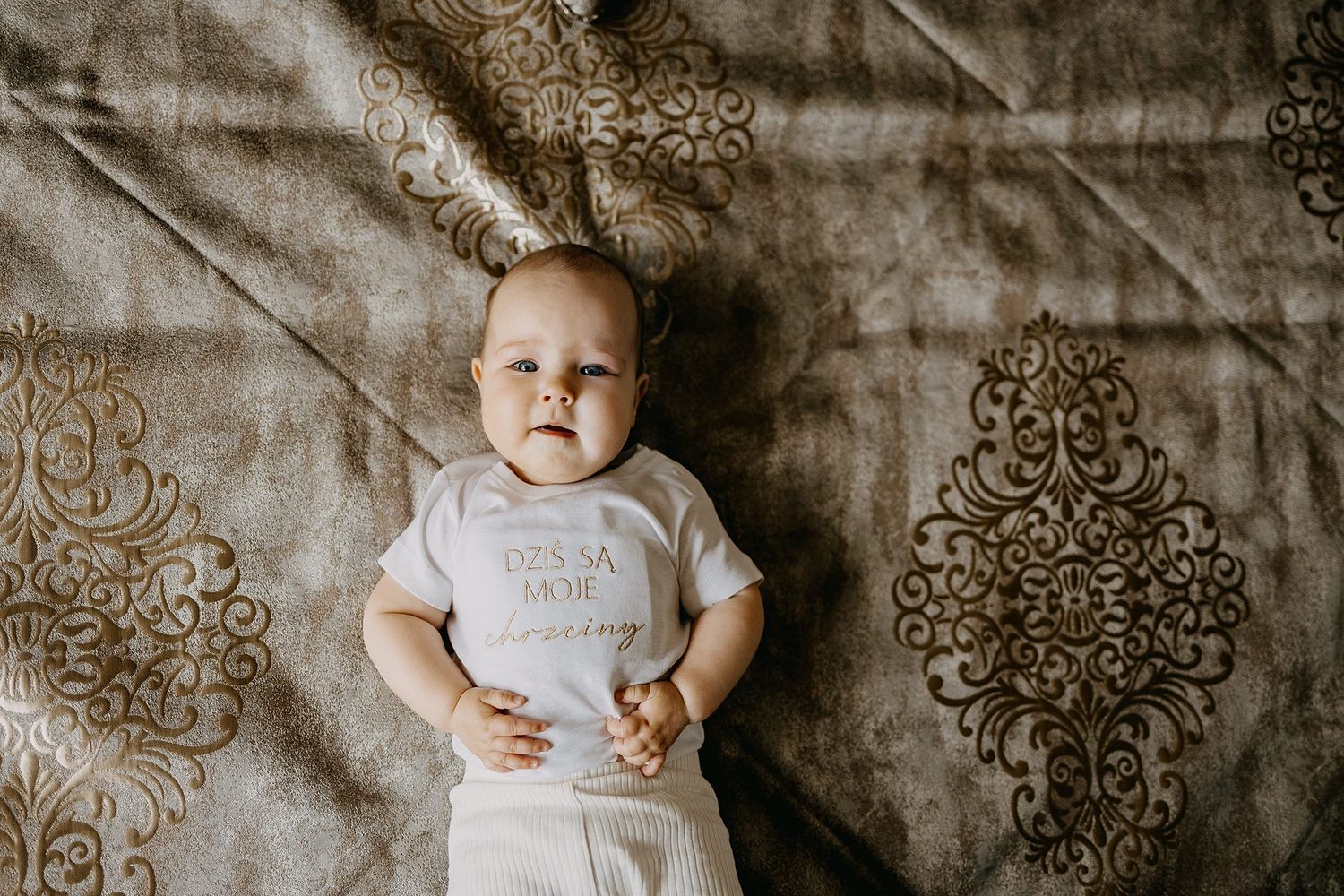 Adorable baby lying on decorative brown fabric wearing white bodysuit at christening celebration.
