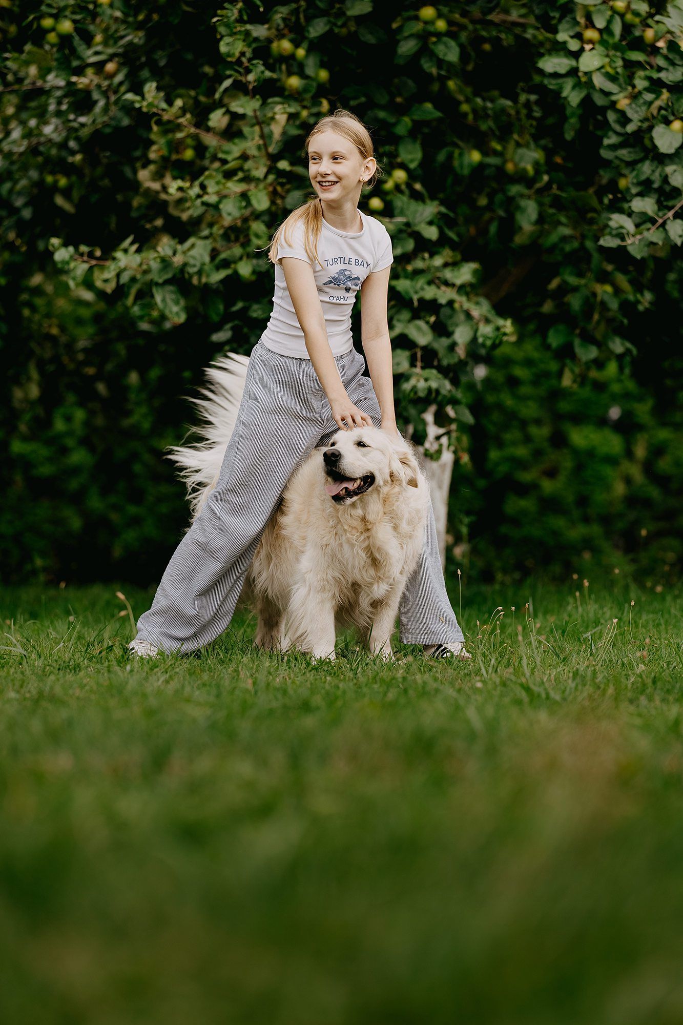 Smiling blonde girl in gray sweatpants playing with a golden retriever on green grass.