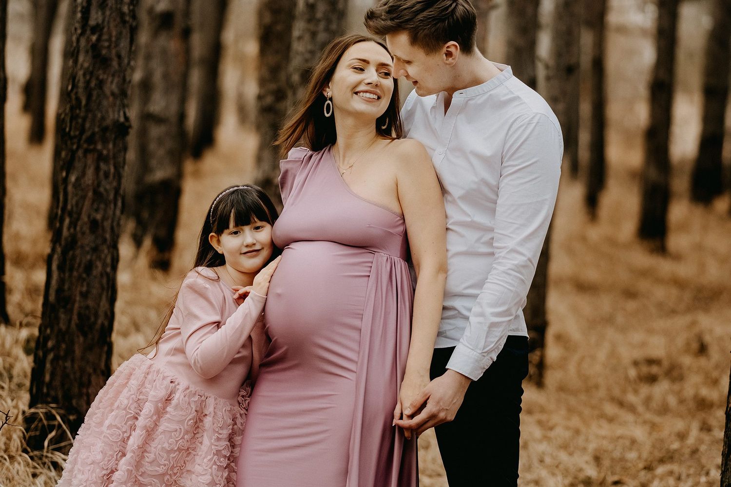 Happy pregnant woman in pink dress with partner and young daughter in autumn forest setting.