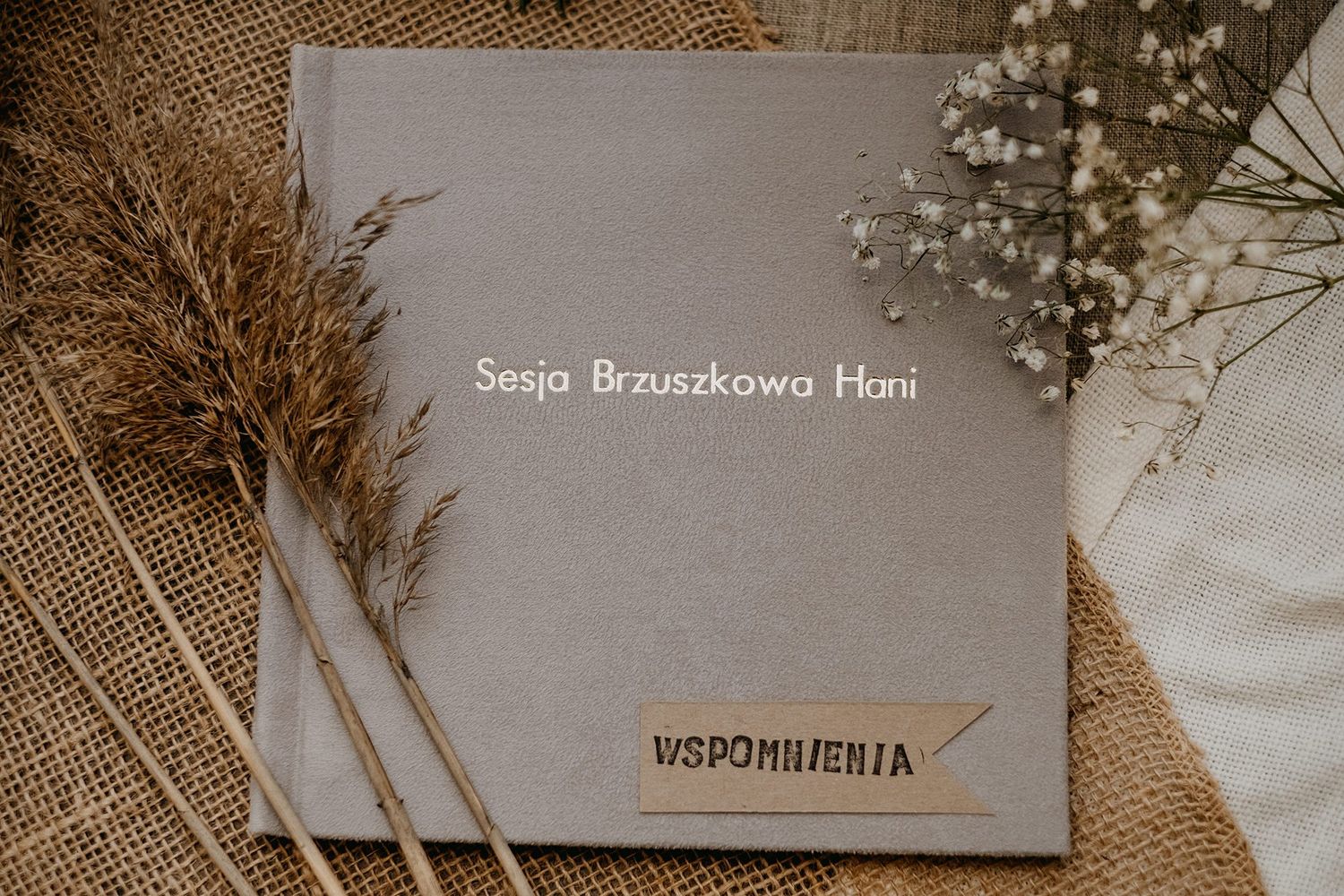 Gray linen photo album cover reading Sesja Brzuszkowa Hani with dried pampas grass and wooden Wspomnienia tag.
