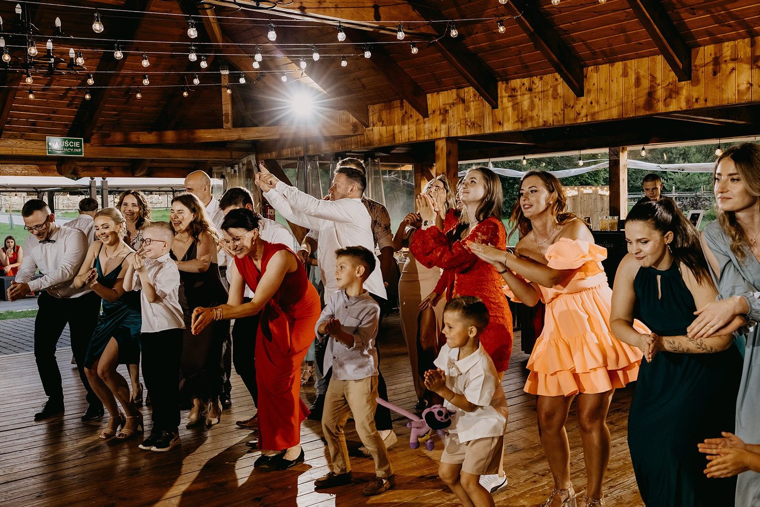 Joyful wedding guests dancing together on a wooden dance floor under warm string lights.