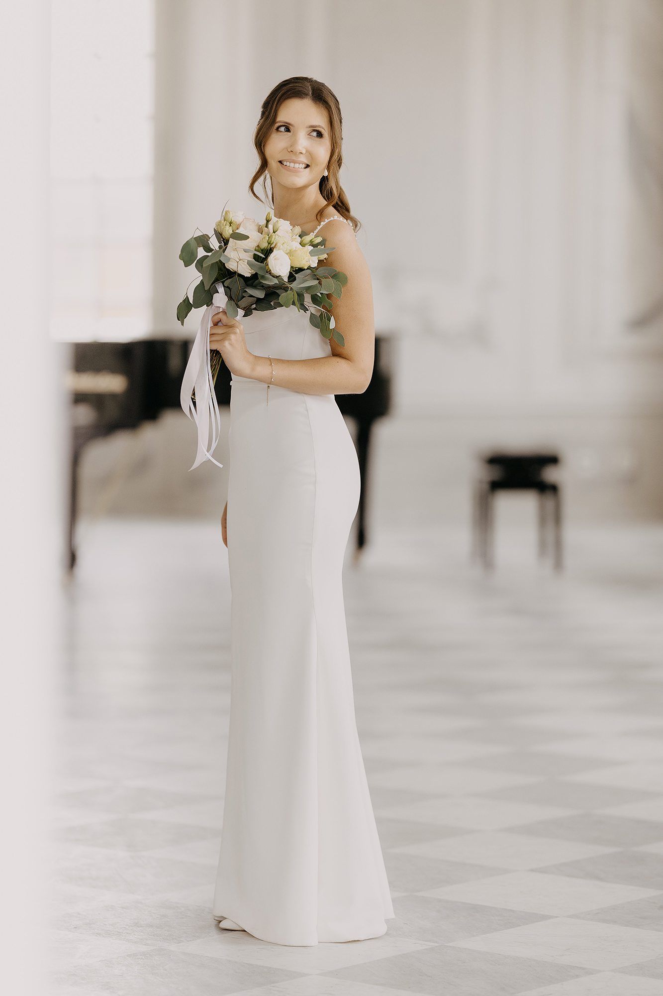 Elegant bride in a white fitted gown holding a bouquet of white roses and greenery in a bright hall.