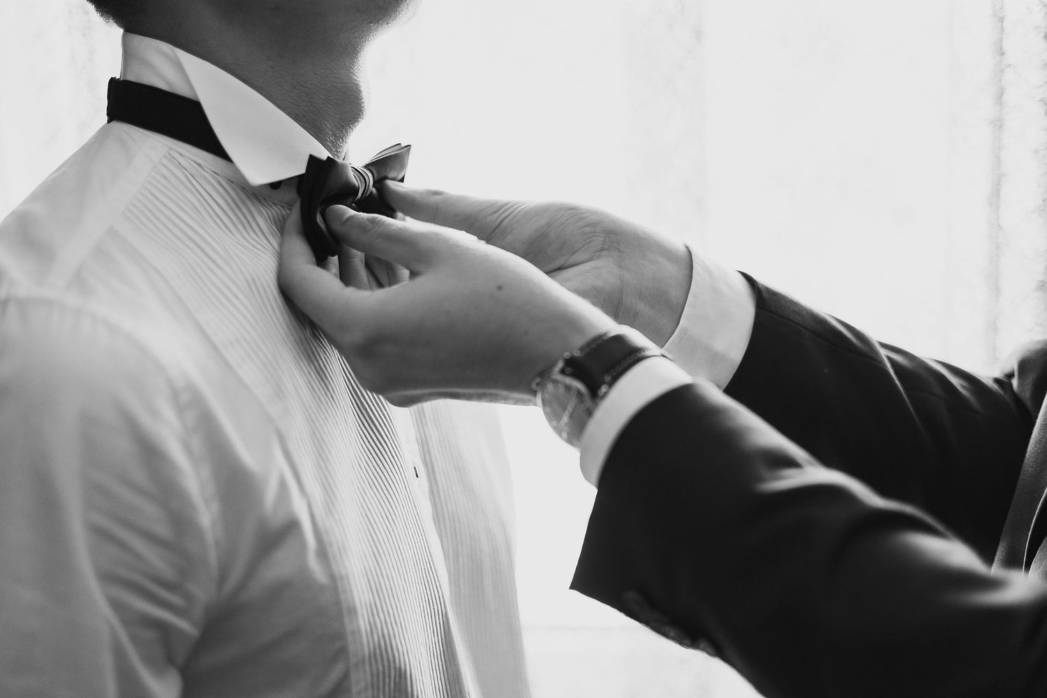 Black and white close-up of groomsman adjusting groom's bow tie before the wedding ceremony.