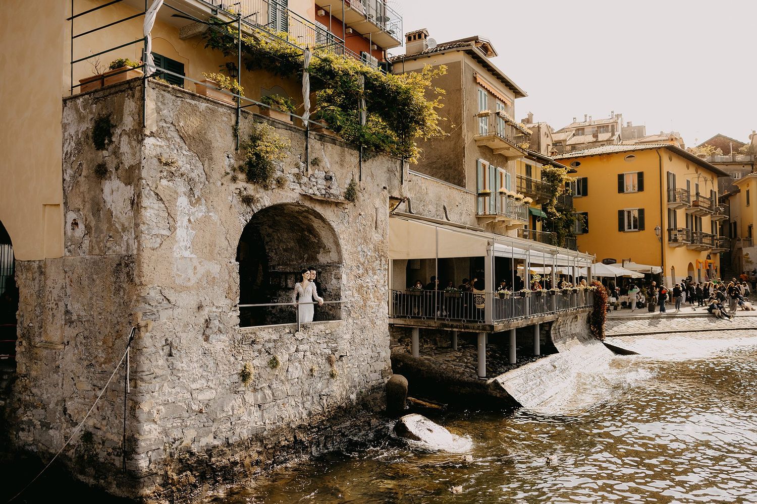 Bride in white gown stands in rustic stone archway beside Italian lakeside village waterfront.