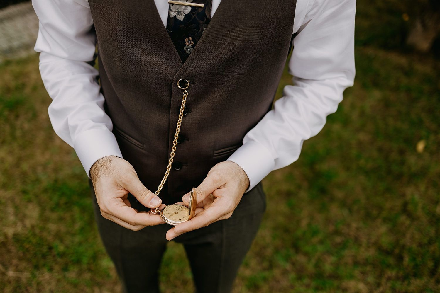 Groom holding a vintage gold pocket watch with chain, wearing a dark brown vest and white shirt.