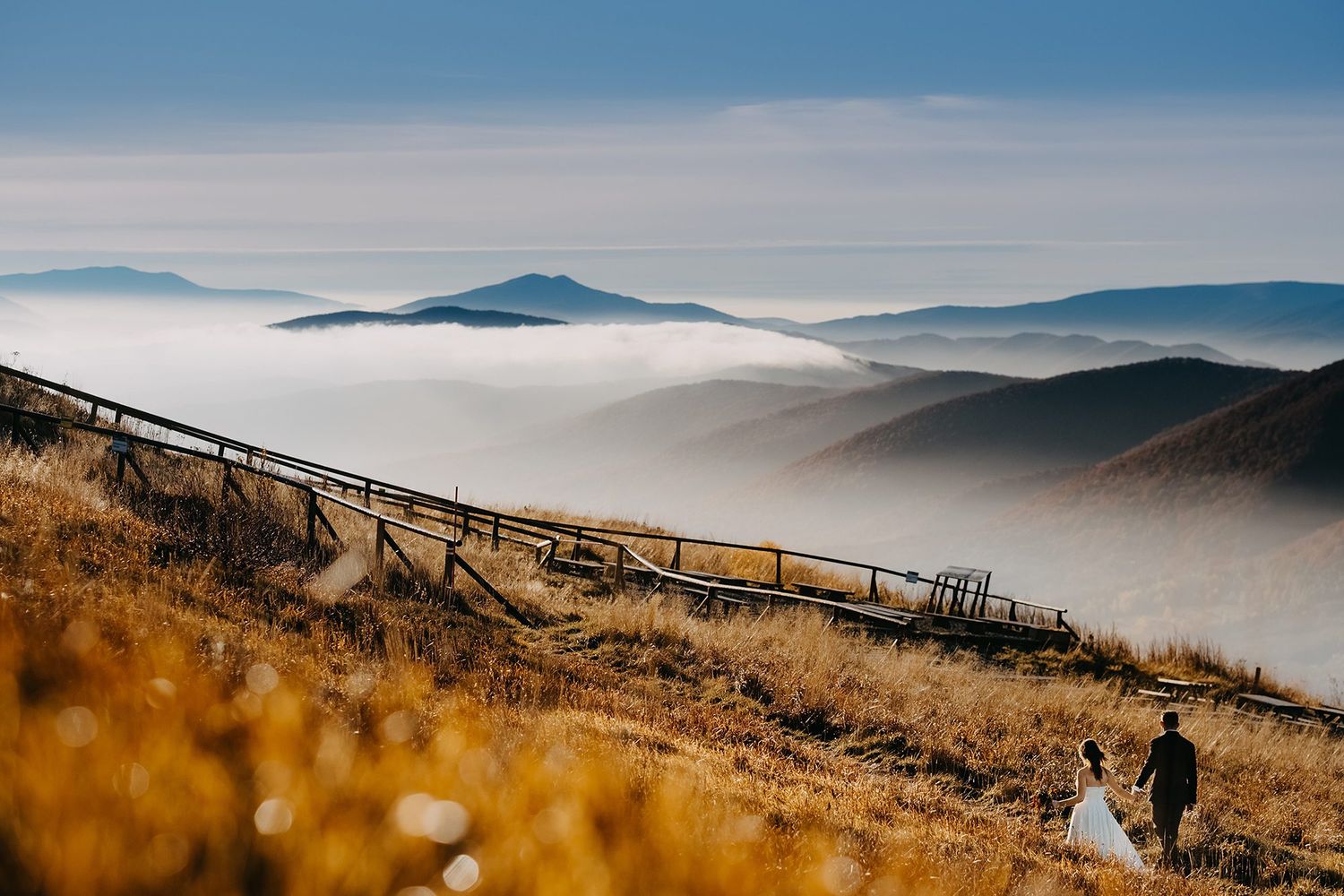 Bride and groom walking on a misty mountain ridge with golden grass and fog-filled valleys below.