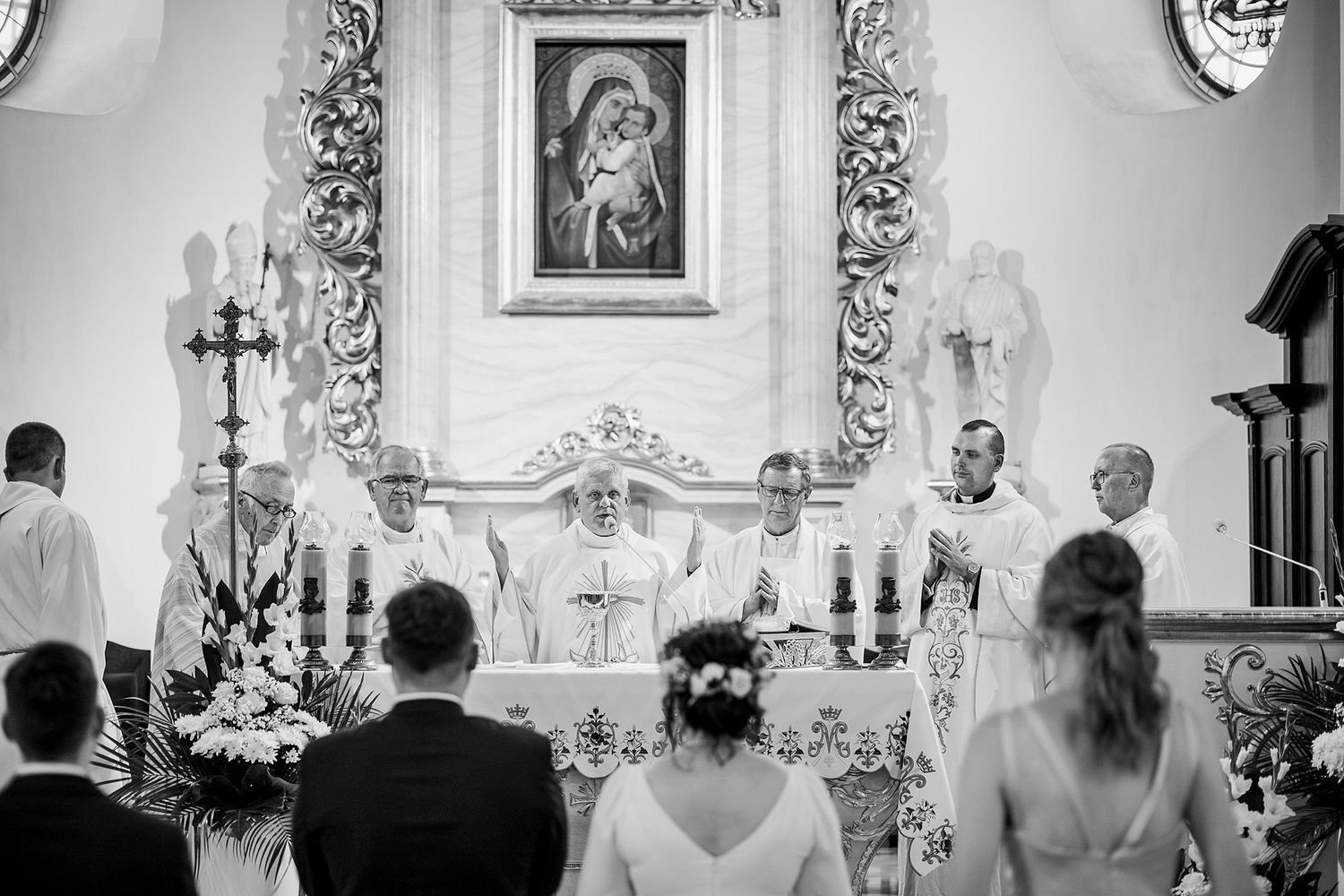 Black and white photo of couple kneeling before priest at ornate church altar during wedding ceremony.