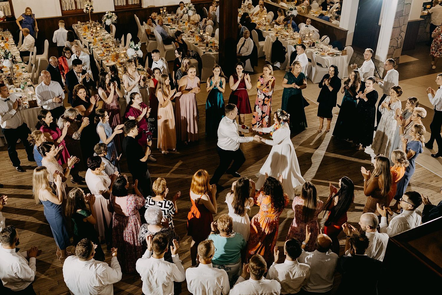 Overhead aerial view of wedding guests dancing together in a large rustic reception hall.