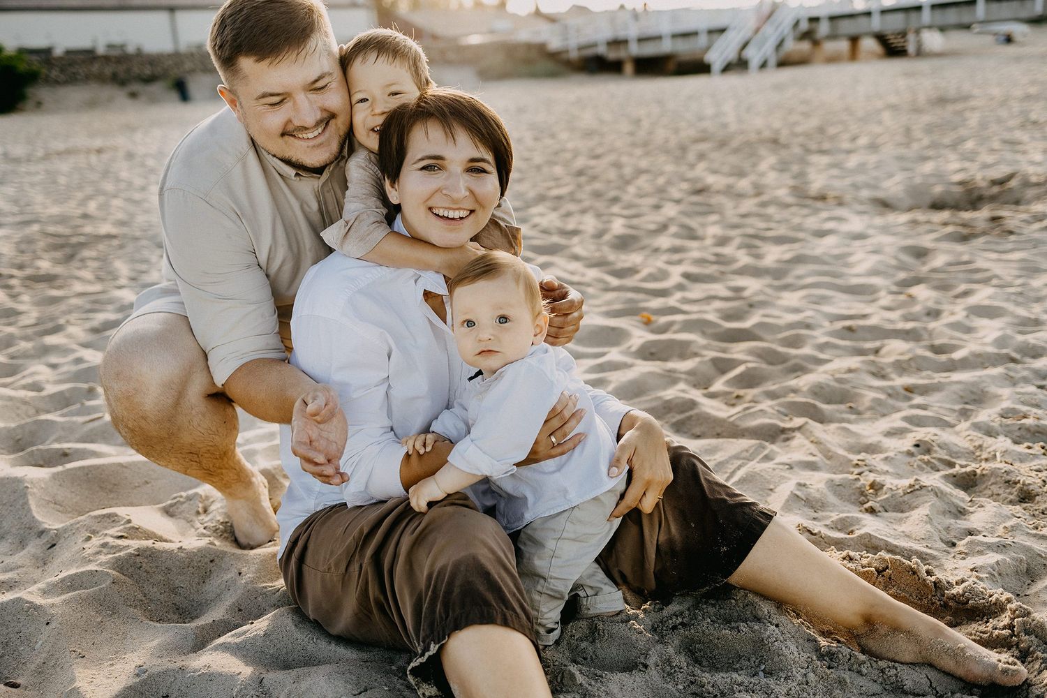 Joyful family of four sitting together on sandy beach during warm sunset, parents hugging children.