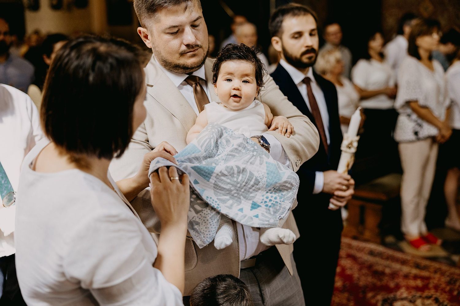 Man in beige suit holding baby wrapped in blue blanket during baptism ceremony.
