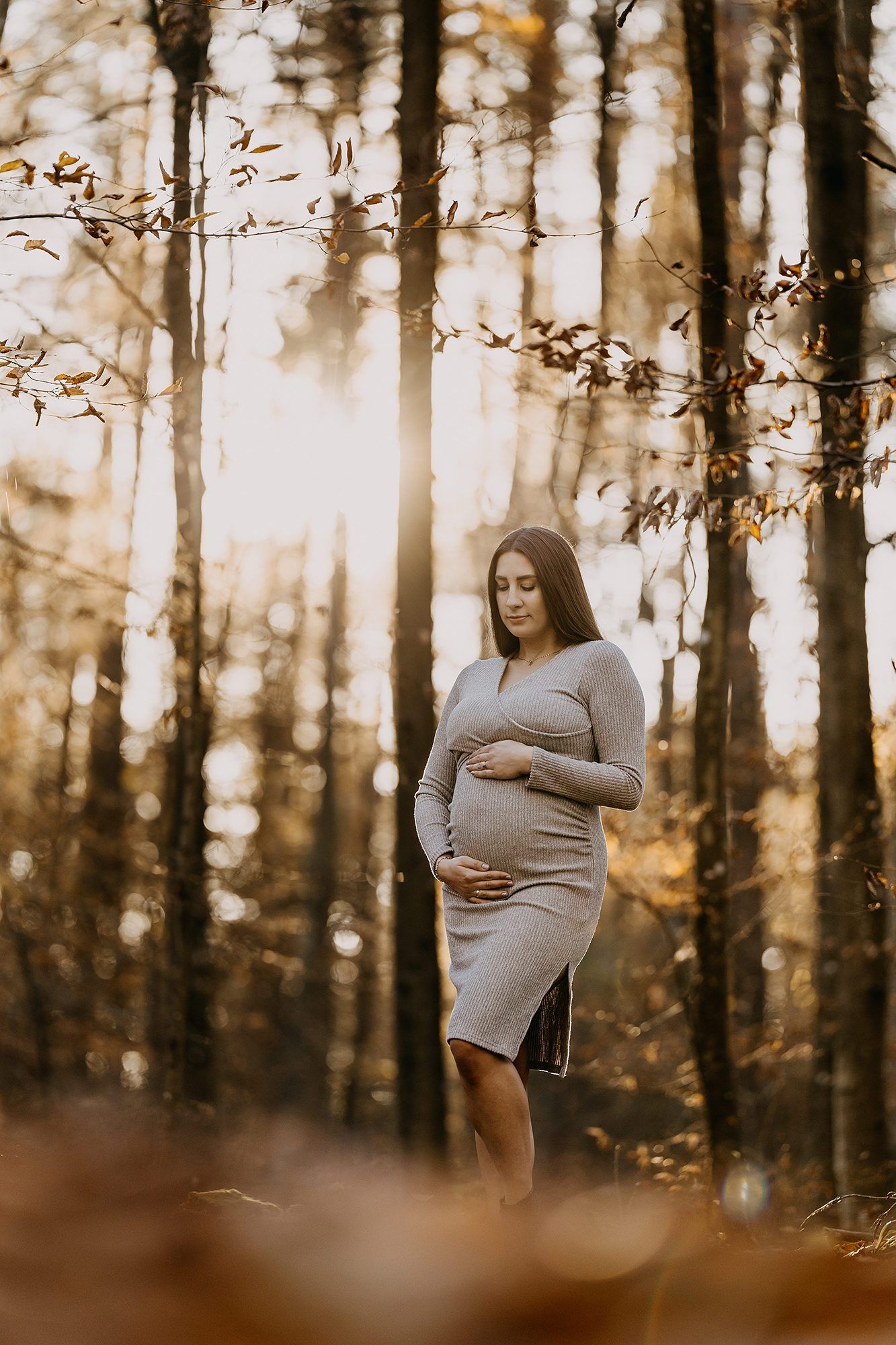 Pregnant woman in grey fitted dress cradling bump in sunlit autumn forest setting.