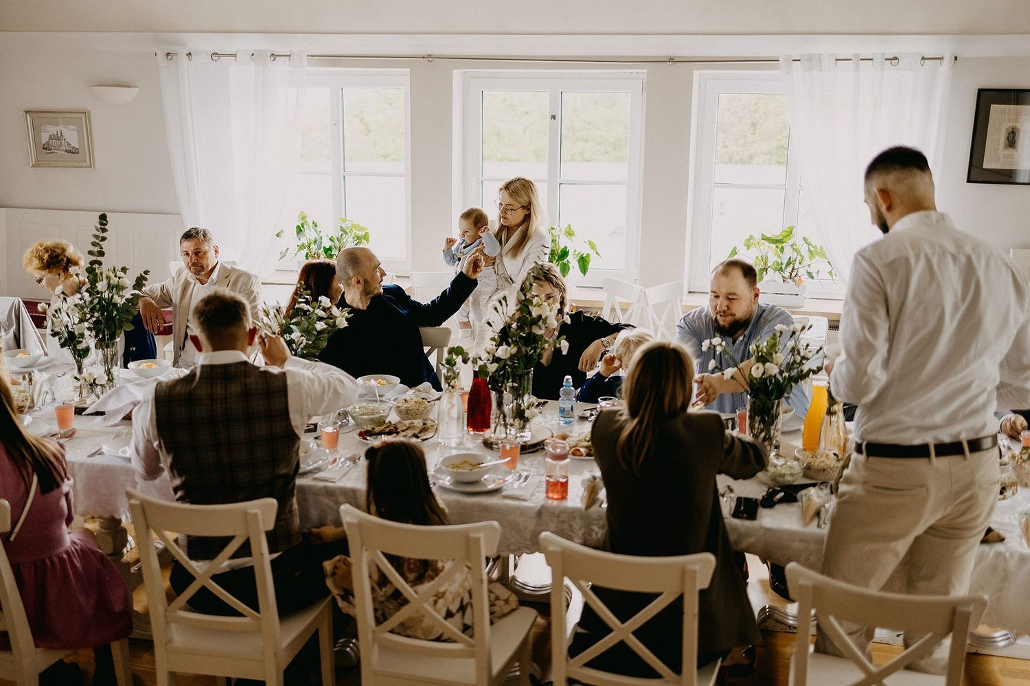 Family and friends gather around decorated table during baptism reception celebration in bright dining room.
