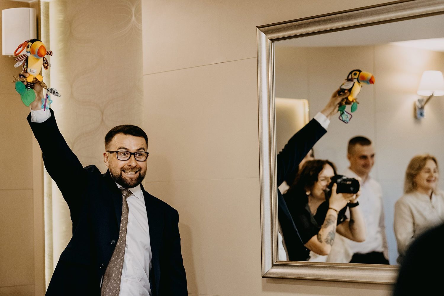 Cheerful man in suit raises colorful baby toys above his head at christening party.