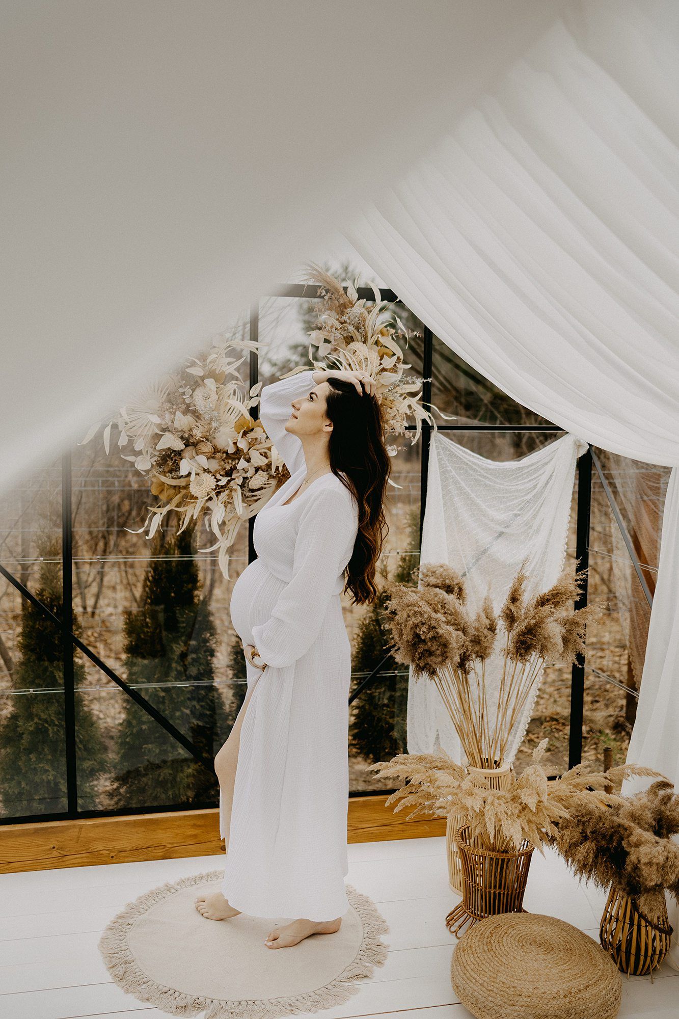 Pregnant woman in white dress holding flower crown beside pampas grass decor in greenhouse studio.