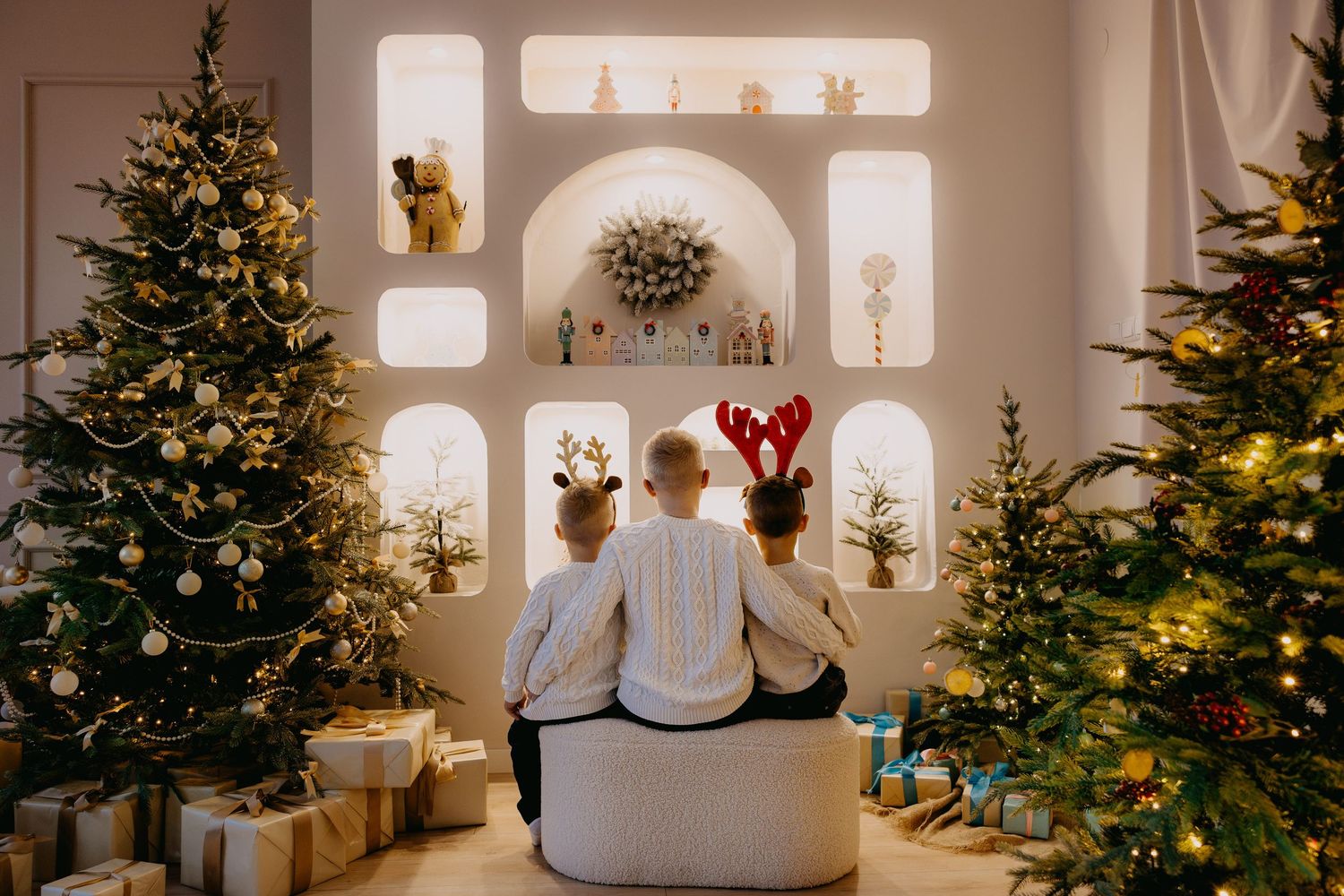 Family of three in white sweaters, two wearing reindeer antlers, sit on a bench between decorated Christmas trees in a cozy festive room.