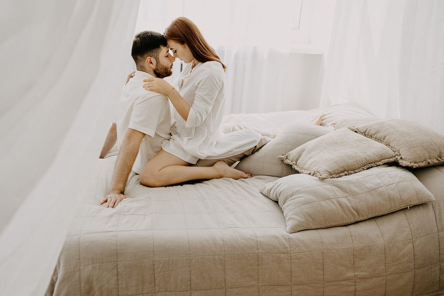 A couple in white robes sitting closely together on a neutral linen bed, sharing an intimate moment.