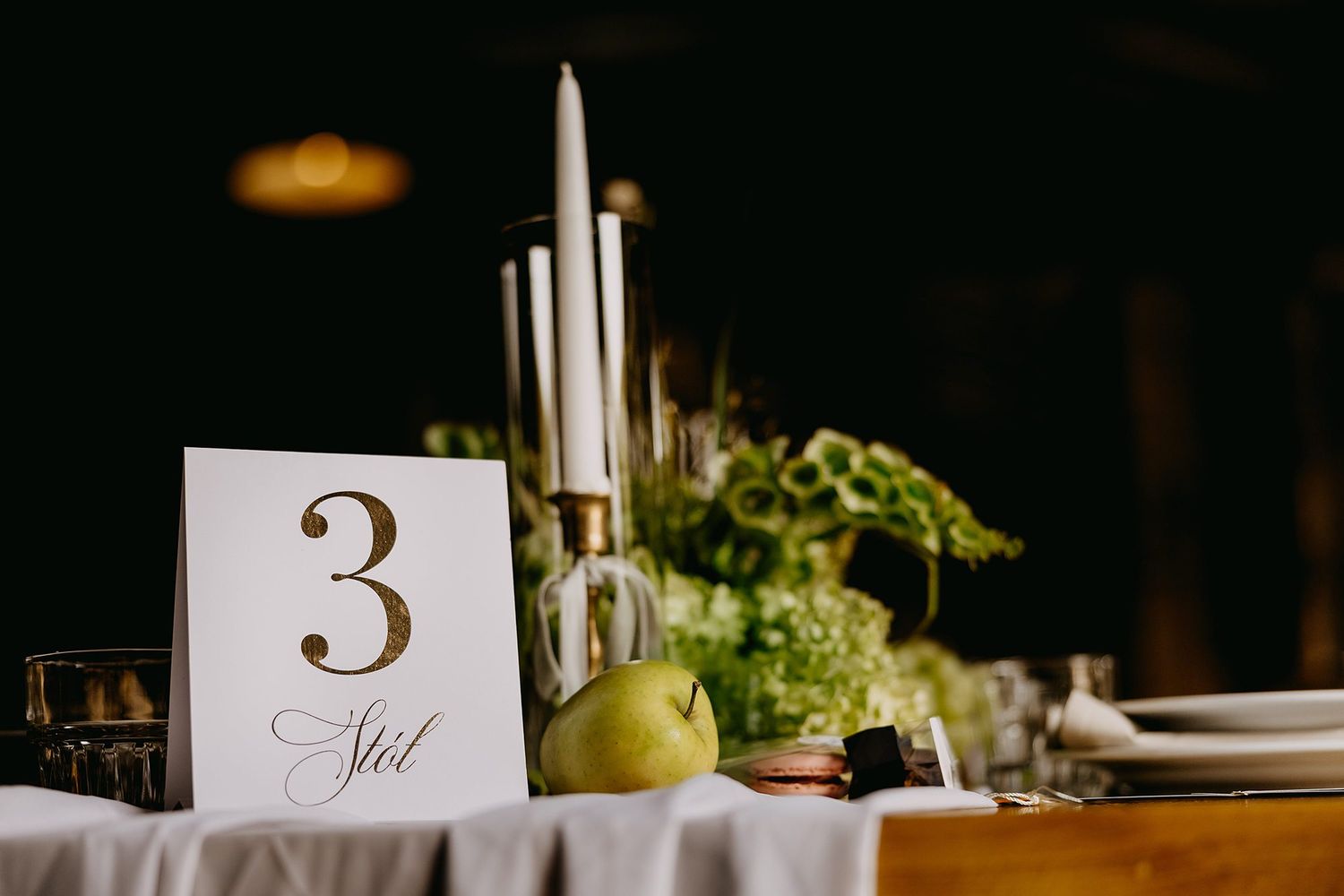 Elegant wedding table number 3 with gold calligraphy, candles, green apple, and floral centerpiece on white linen.