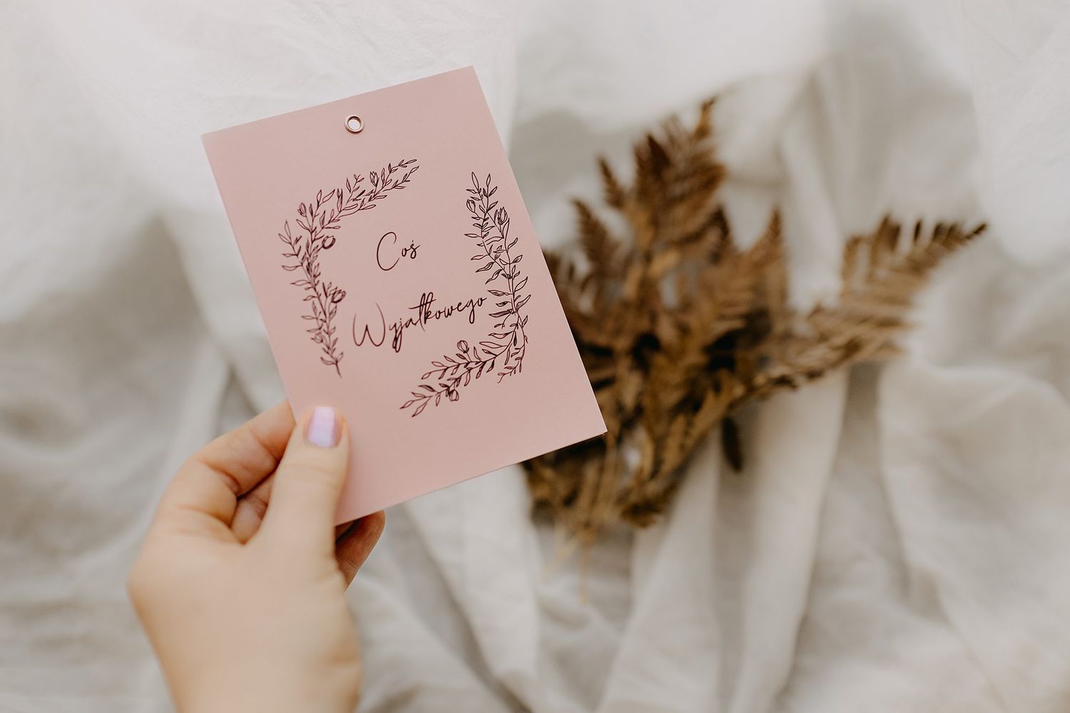 Hand holding pink gift tag with wreath illustration and Cos Wyjatkowego text near dried fern leaves.