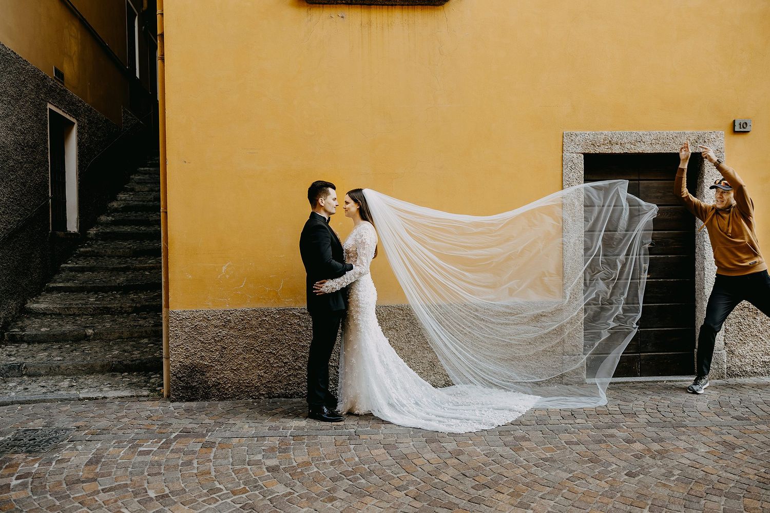 Bride and groom embracing while assistant tosses flowing veil against rustic yellow stone wall.