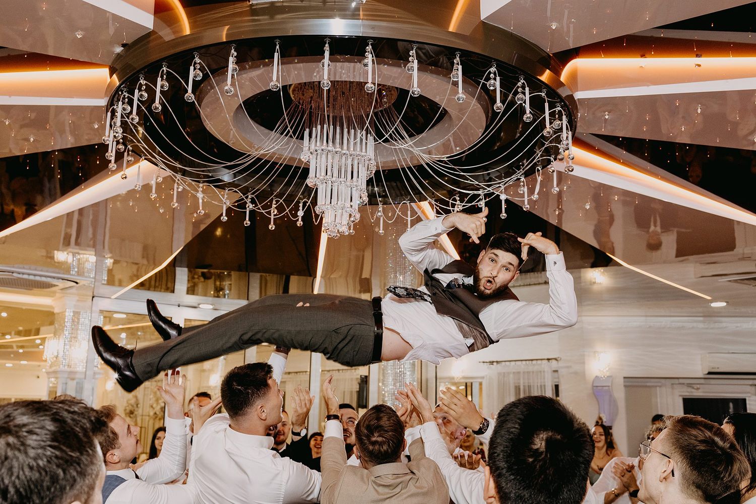 Groom is tossed high into the air by groomsmen beneath a chandelier at wedding reception.