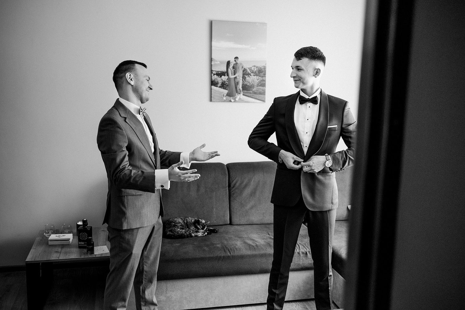 Black and white photo of groom in tuxedo chatting with groomsman while getting ready.
