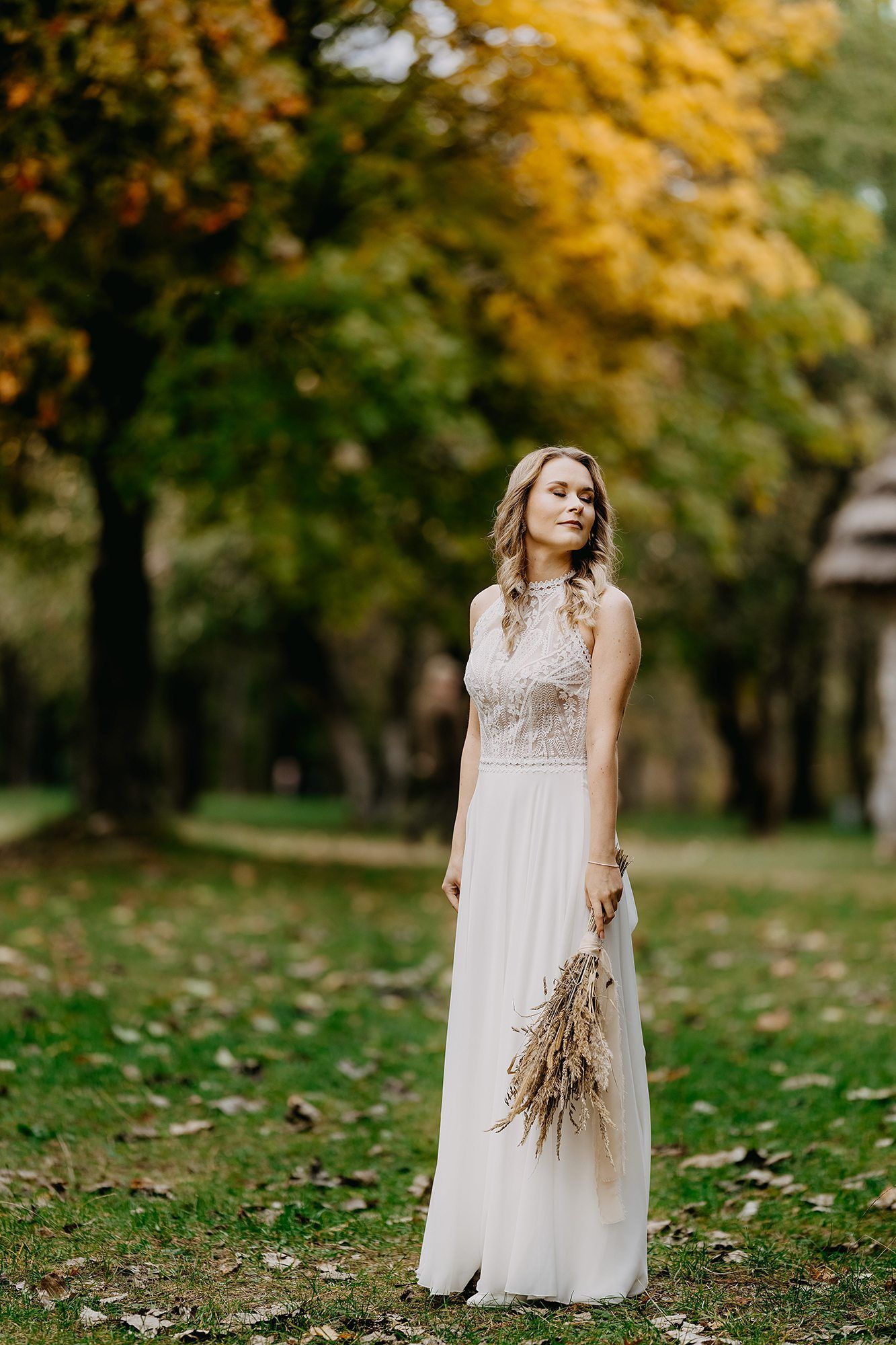 Elegant bride in white gown holding a dried bouquet in a colorful autumn park.