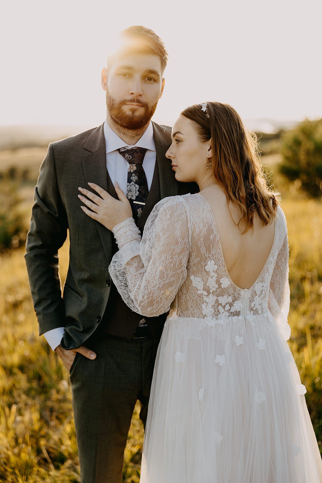 Bride in lace gown and groom in dark suit embracing at golden sunset in a field.