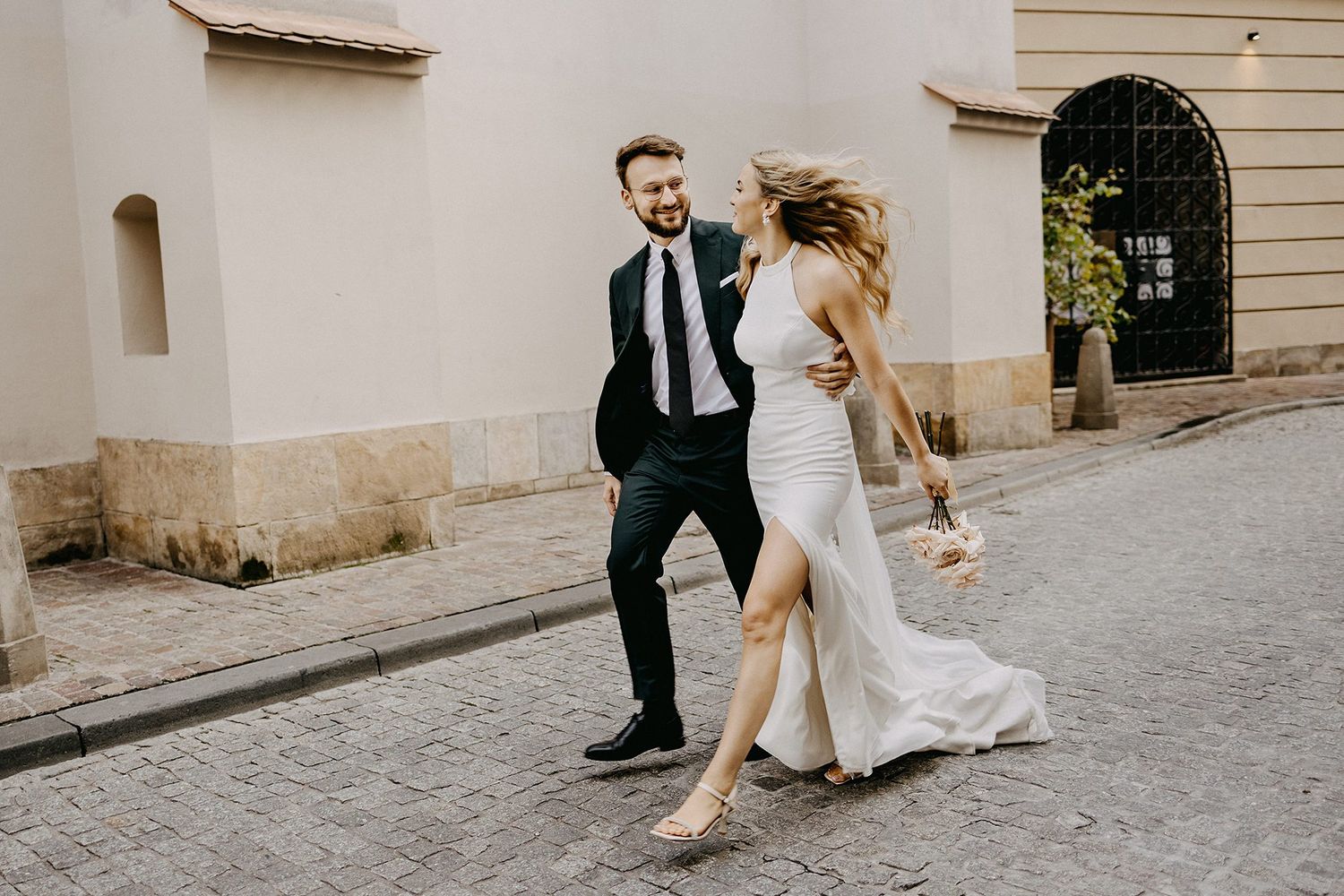 Joyful bride and groom running and laughing on cobblestone street in Europe.
