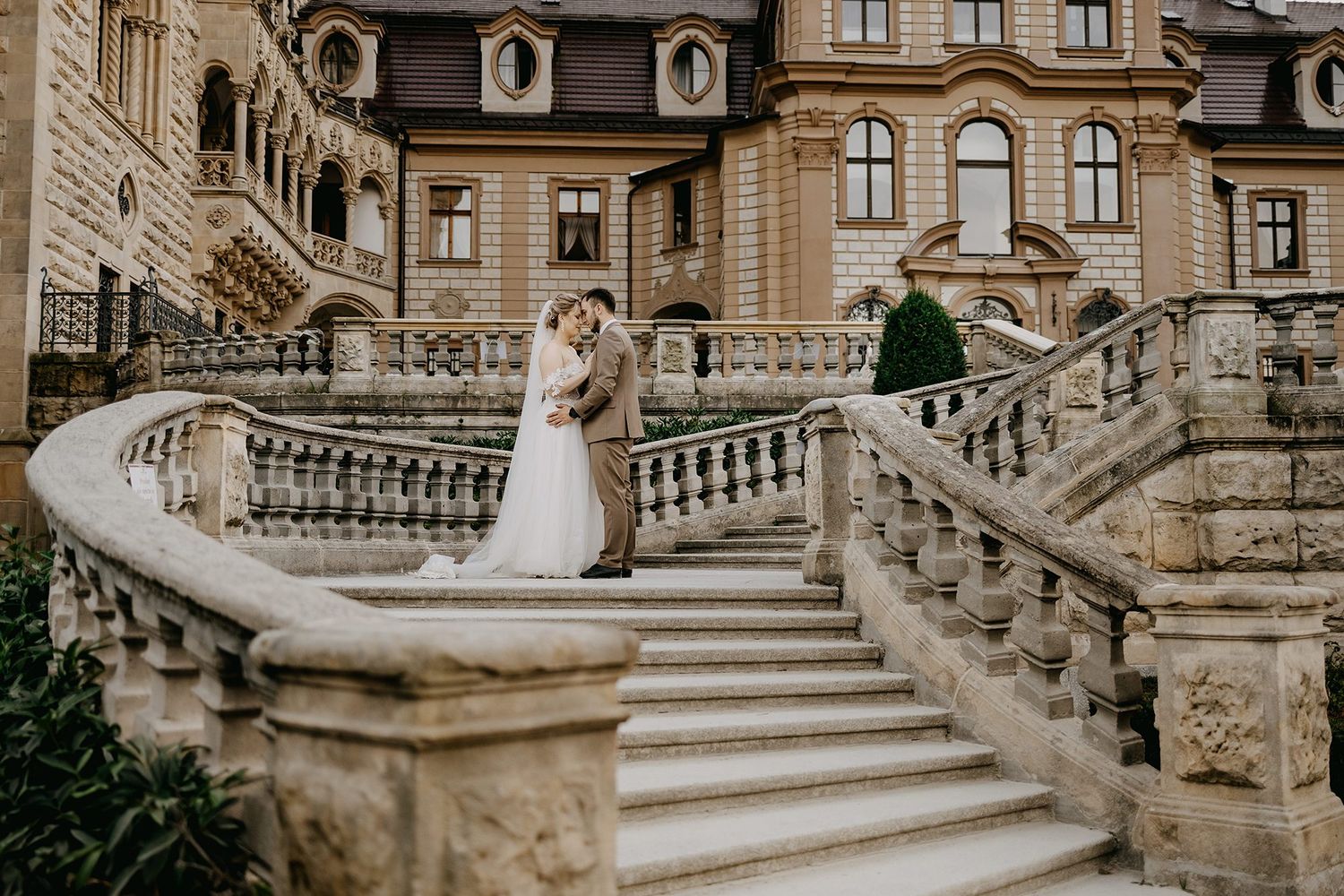 Elegant bride and groom embracing on grand stone staircase of castle.