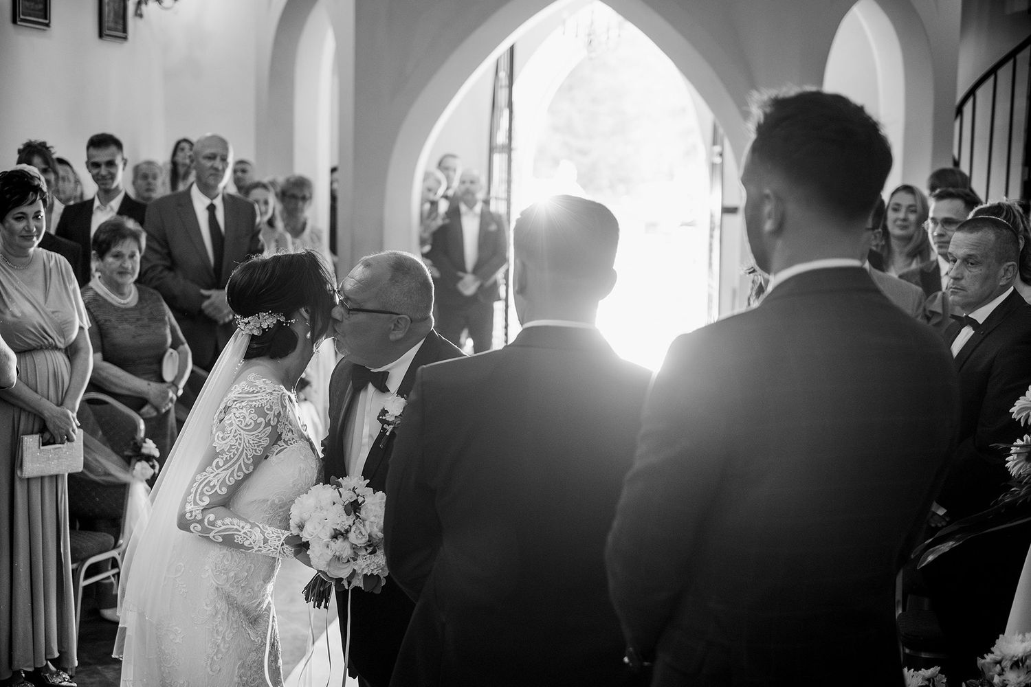 Black and white photo of bride receiving kiss from father before groom at sunlit church ceremony.