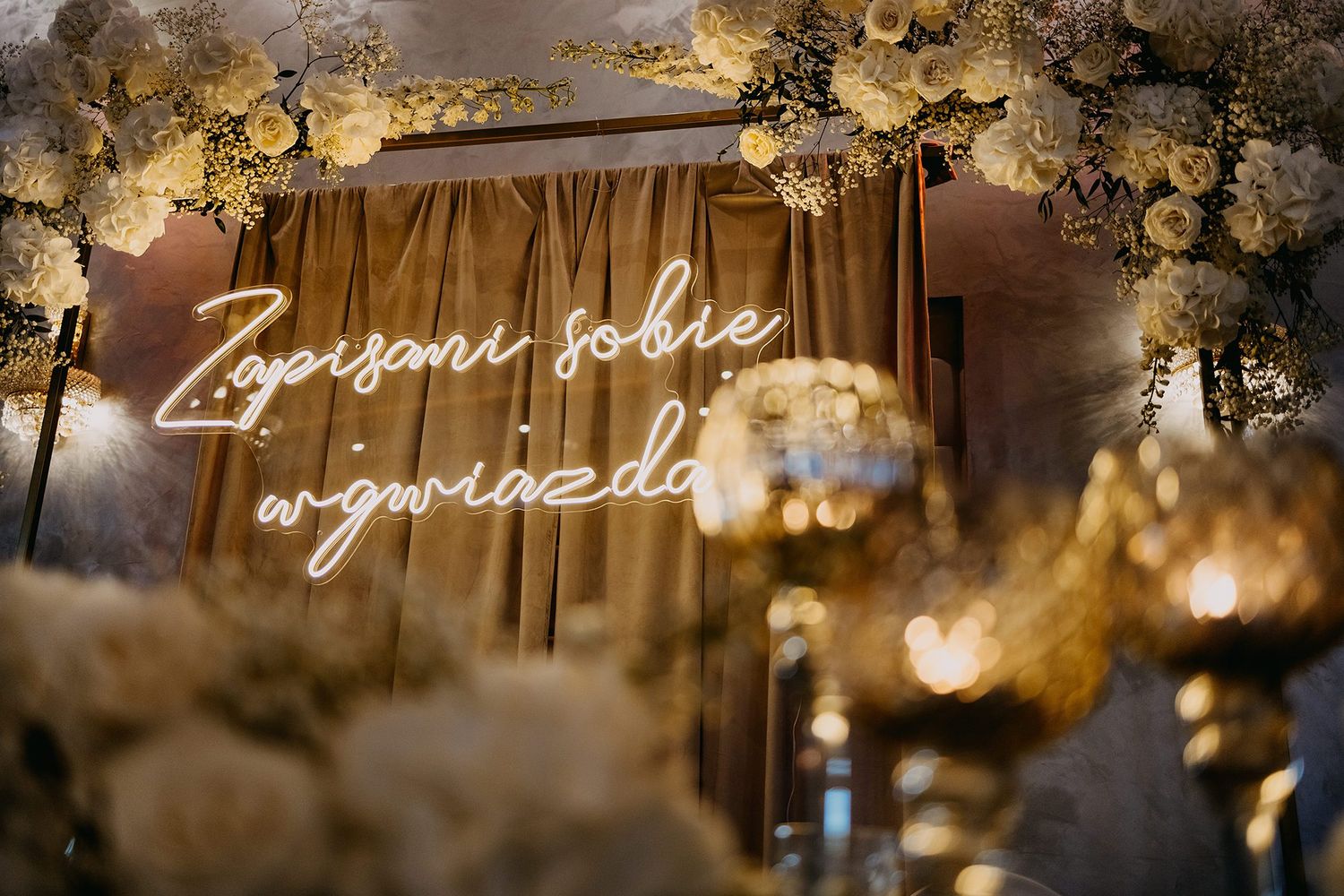 Neon sign reading Zapisani sobie w gwiazdach surrounded by white floral wedding decor.