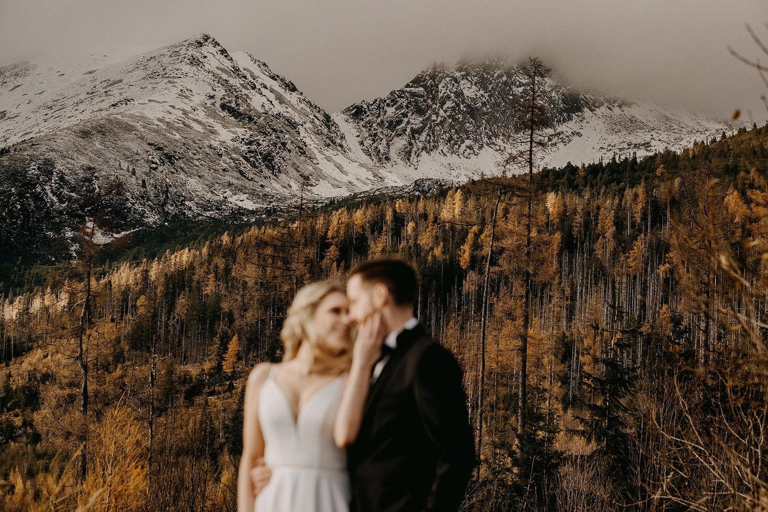 Couple share a kiss with blurred foreground and dramatic snow-covered mountain backdrop.