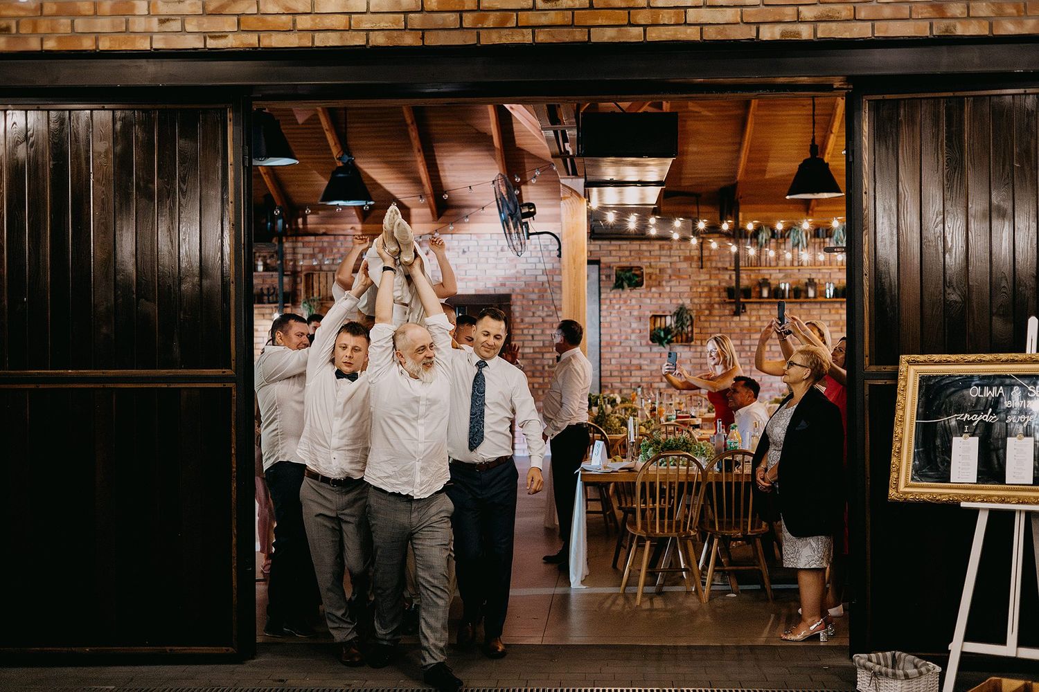 Wedding guests cheerfully entering a rustic brick venue through large wooden doors with string lights inside.