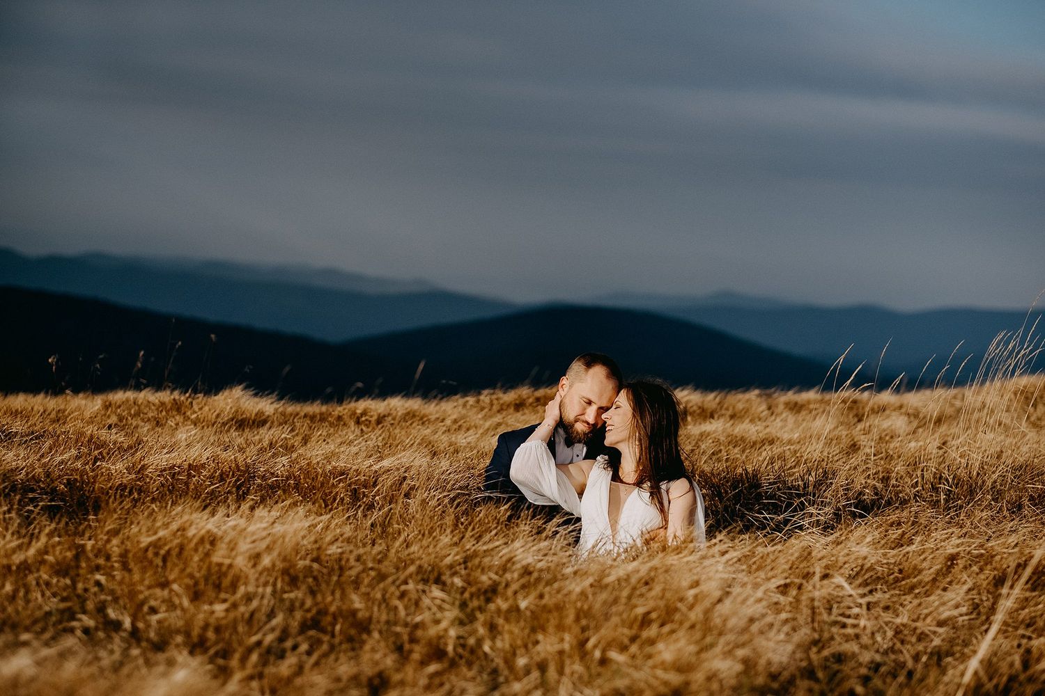 Bride and groom lying together in golden grass on a mountain top under moody blue skies.