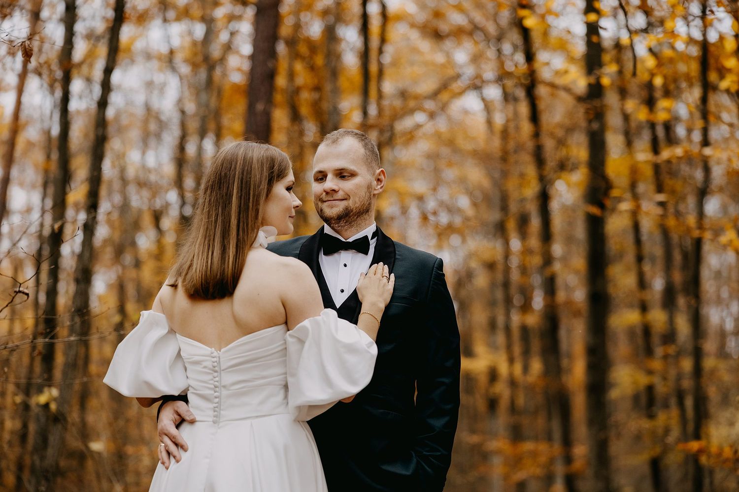 Smiling groom in tuxedo and bride in off-shoulder gown pose in golden autumn forest.