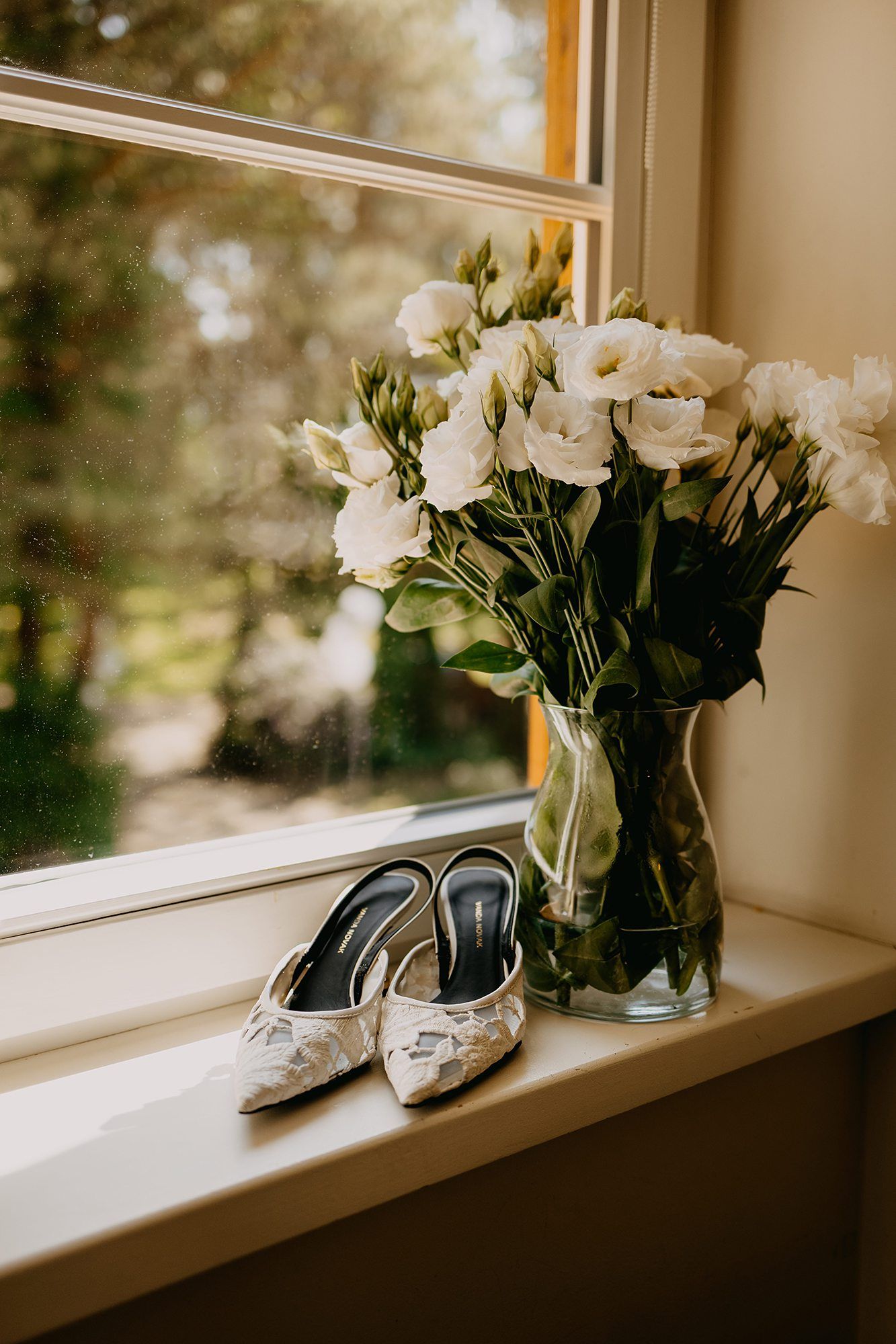 Elegant lace bridal heels on windowsill beside glass vase of white lisianthus flowers.