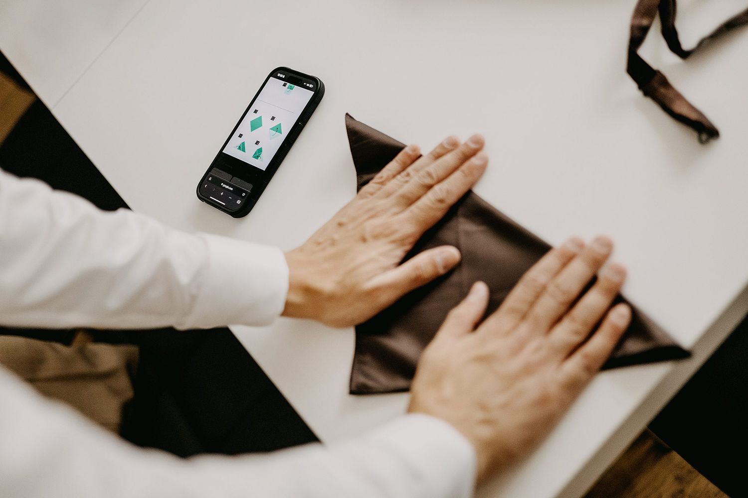 Close-up of hands smoothing fabric on a table beside a smartphone displaying a colorful app.