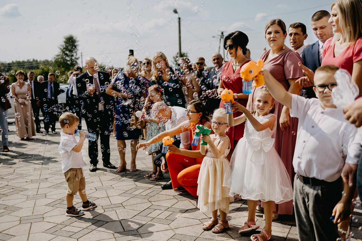 Children and wedding guests celebrating outdoors with balloons and confetti in sunlight.