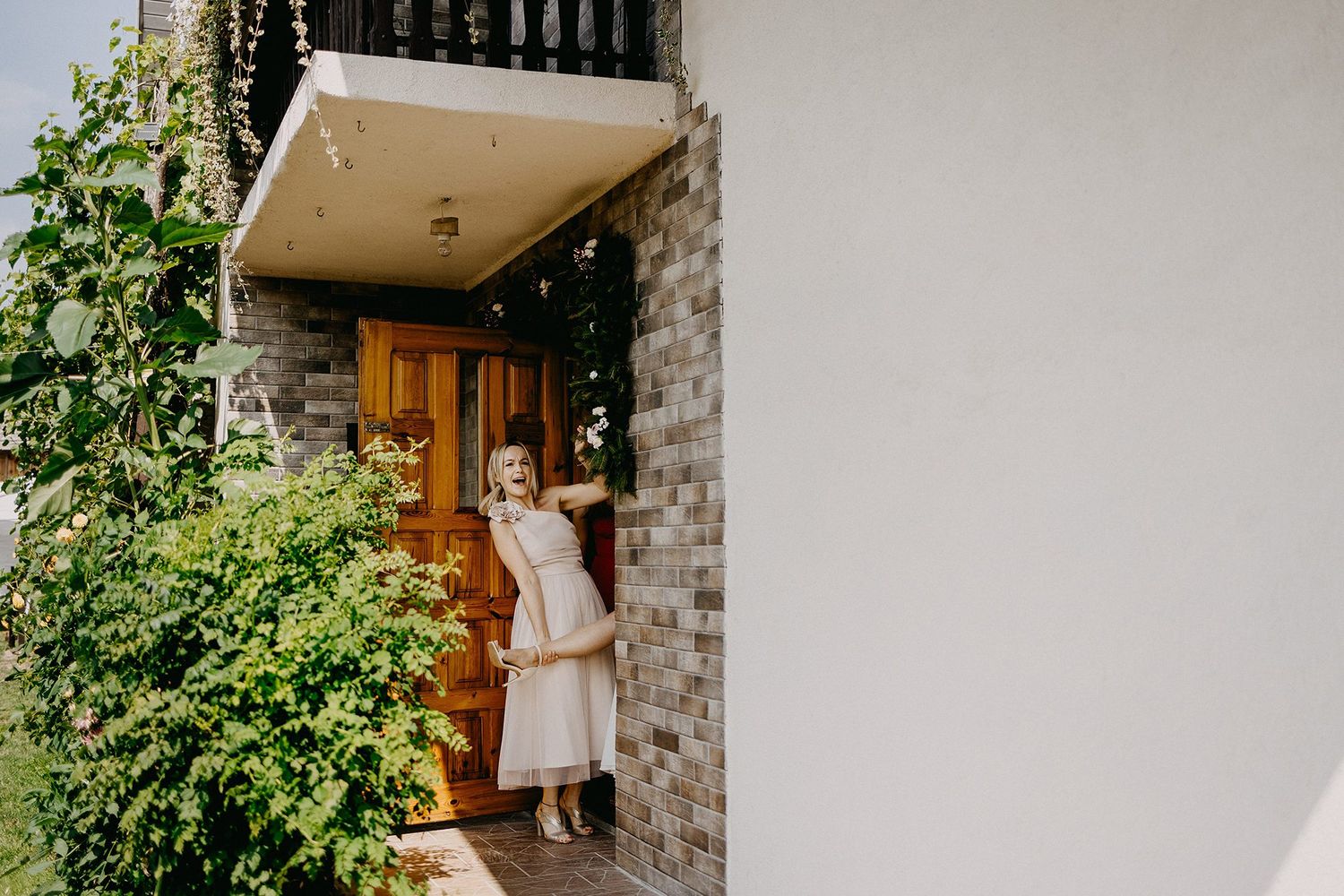 Bride in white dress standing at wooden doorway surrounded by lush green garden foliage.